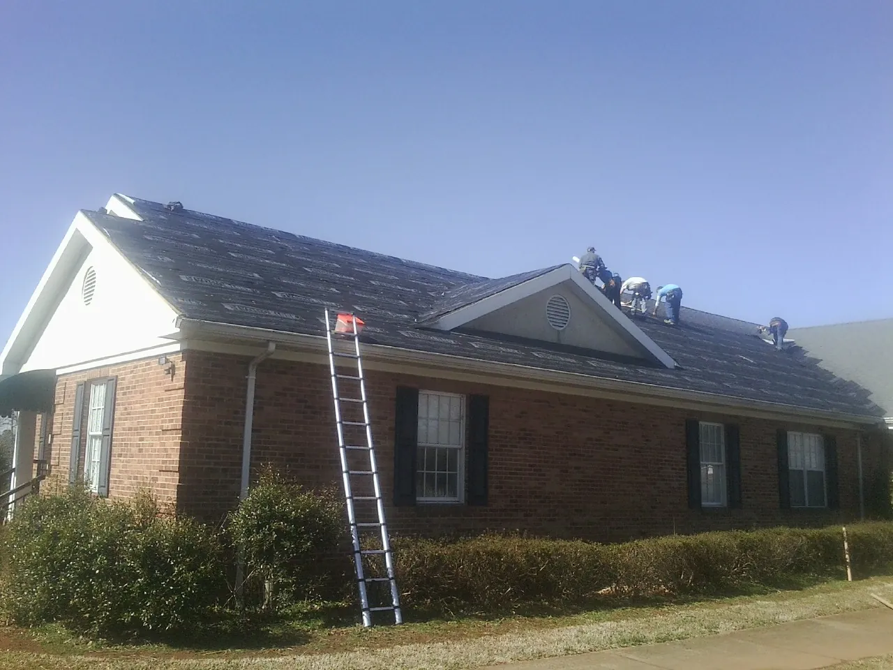 Crew tearing shingles off a brick ranch roof with a ladder set — Metal Roof Repair work by our crew in High Ridge