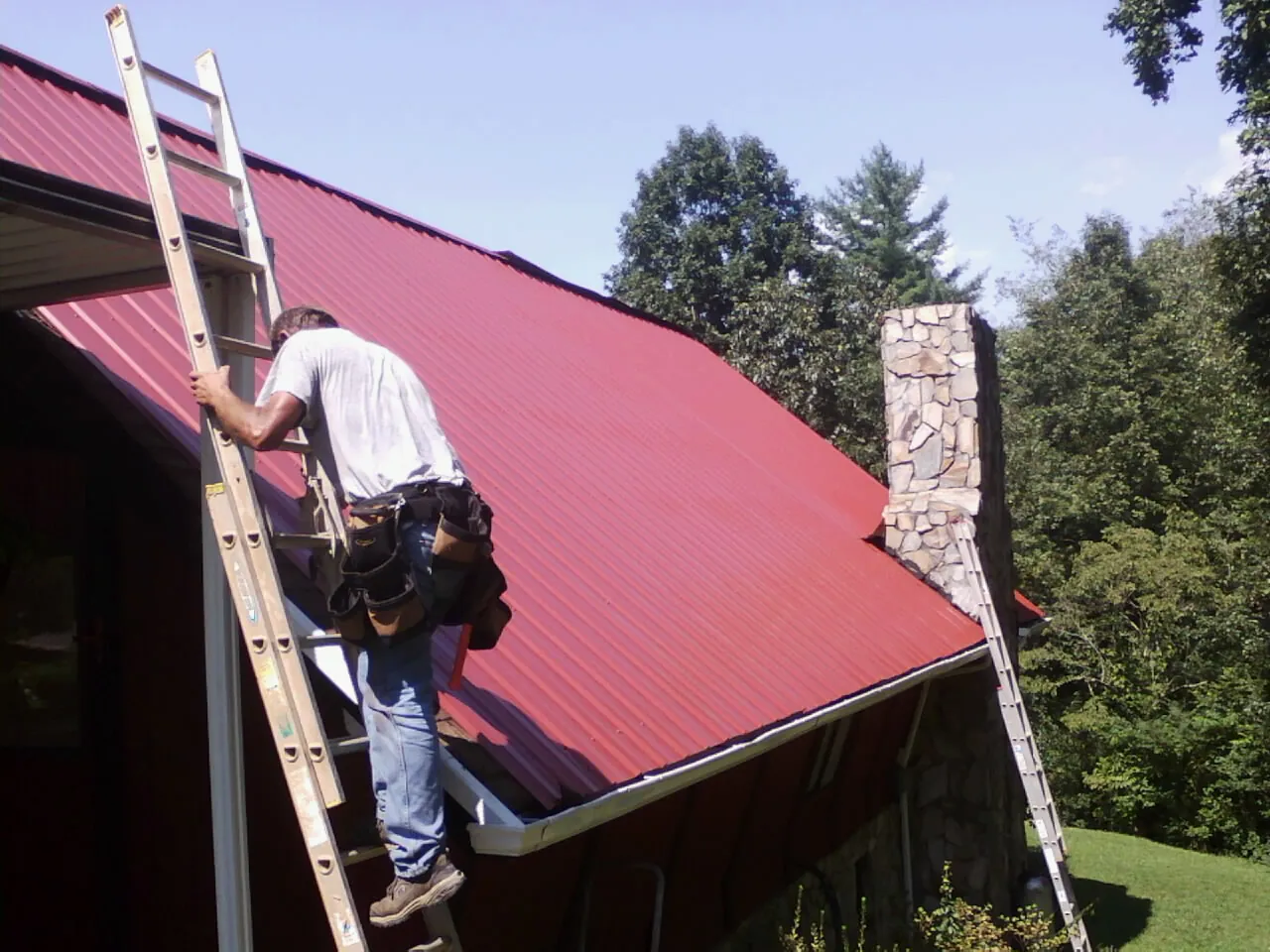 Roofer on a ladder beside a red metal roof and stone chimney — Soffit & Fascia Repair by Roofing Brothers in High Ridge