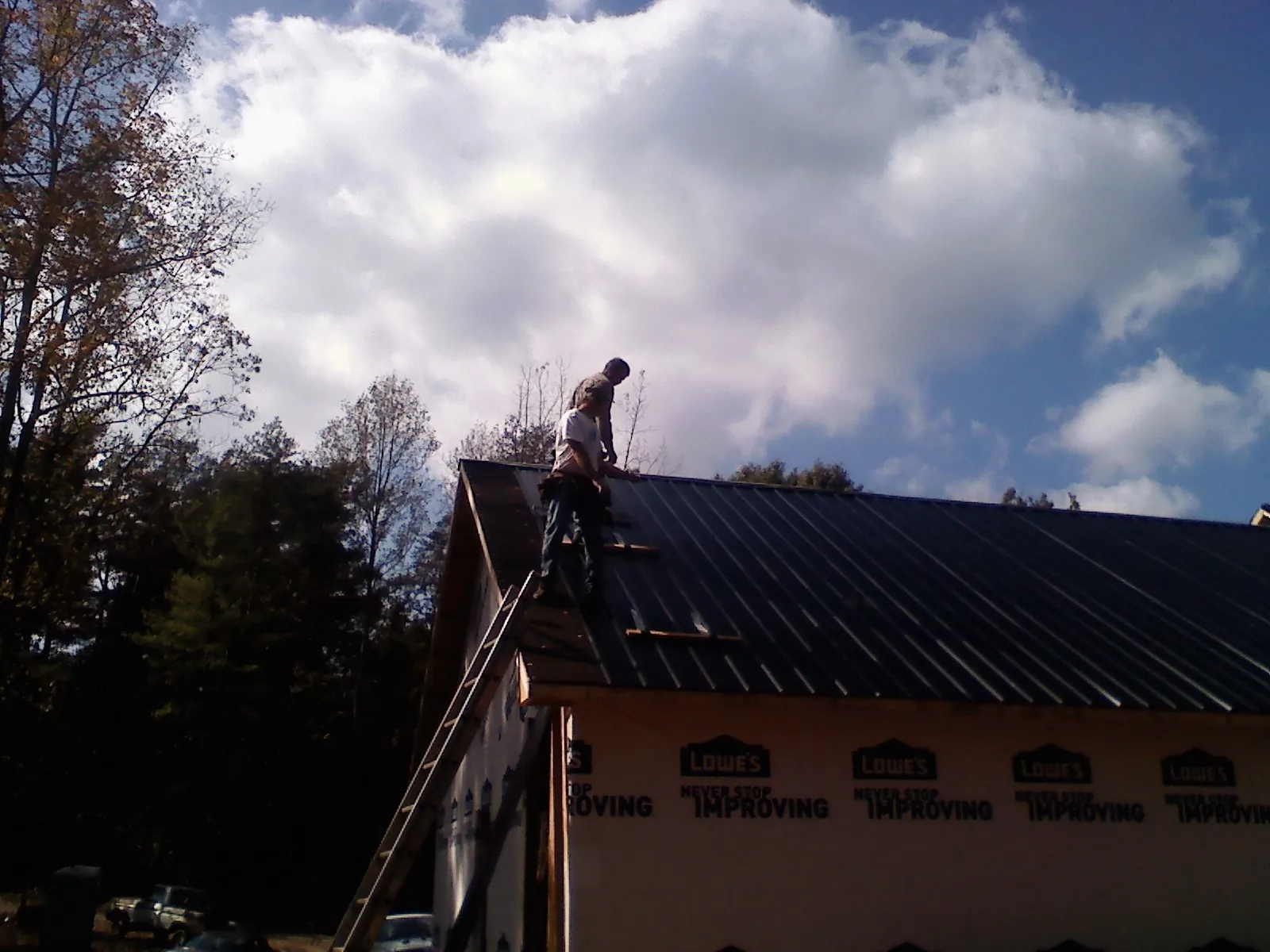 Roofer fastening dark metal panels high against a cloudy sky, from a Roof Inspection job in High Ridge